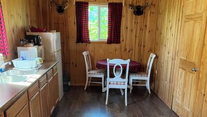 Een houten chalet van binnen, met een keuken, twee witte stoelen, een eettafel en een raam met rode gordijnen.