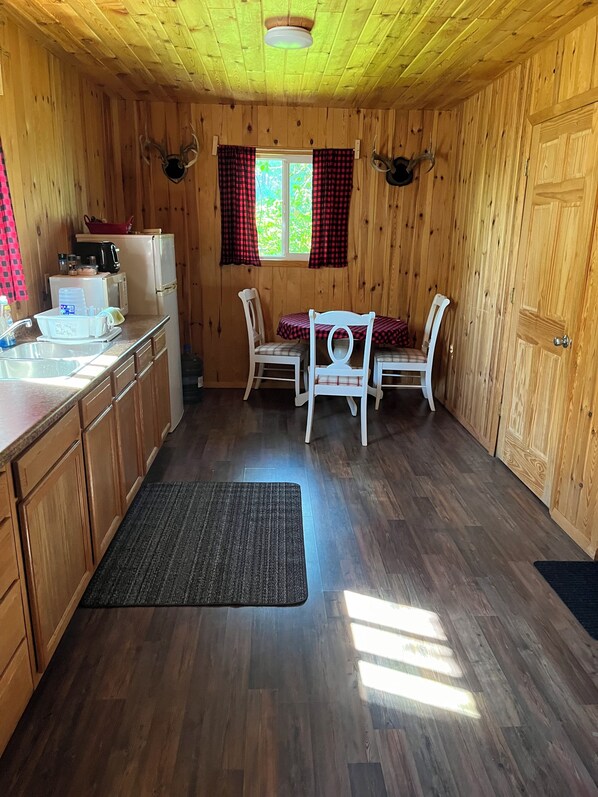 A wooden cabin interior with a kitchen area, two white chairs, a dining table, and a window with red curtains.