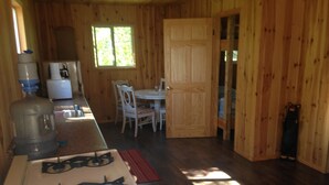 Interieur van een houten chalet met een keukenhoek, inclusief een fornuis, een groot waterdispenser en een koelkast. Er is een eettafel met stoelen en een raam zorgt voor natuurlijk licht.