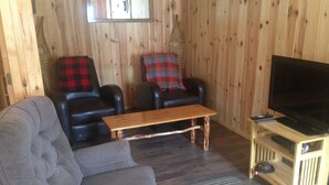 Een gezellige woonkamer met een grijze bank, twee zwarte leren fauteuils met rood-zwarte geruite plaids, een houten salontafel, en een televisie op een houten dressoir.