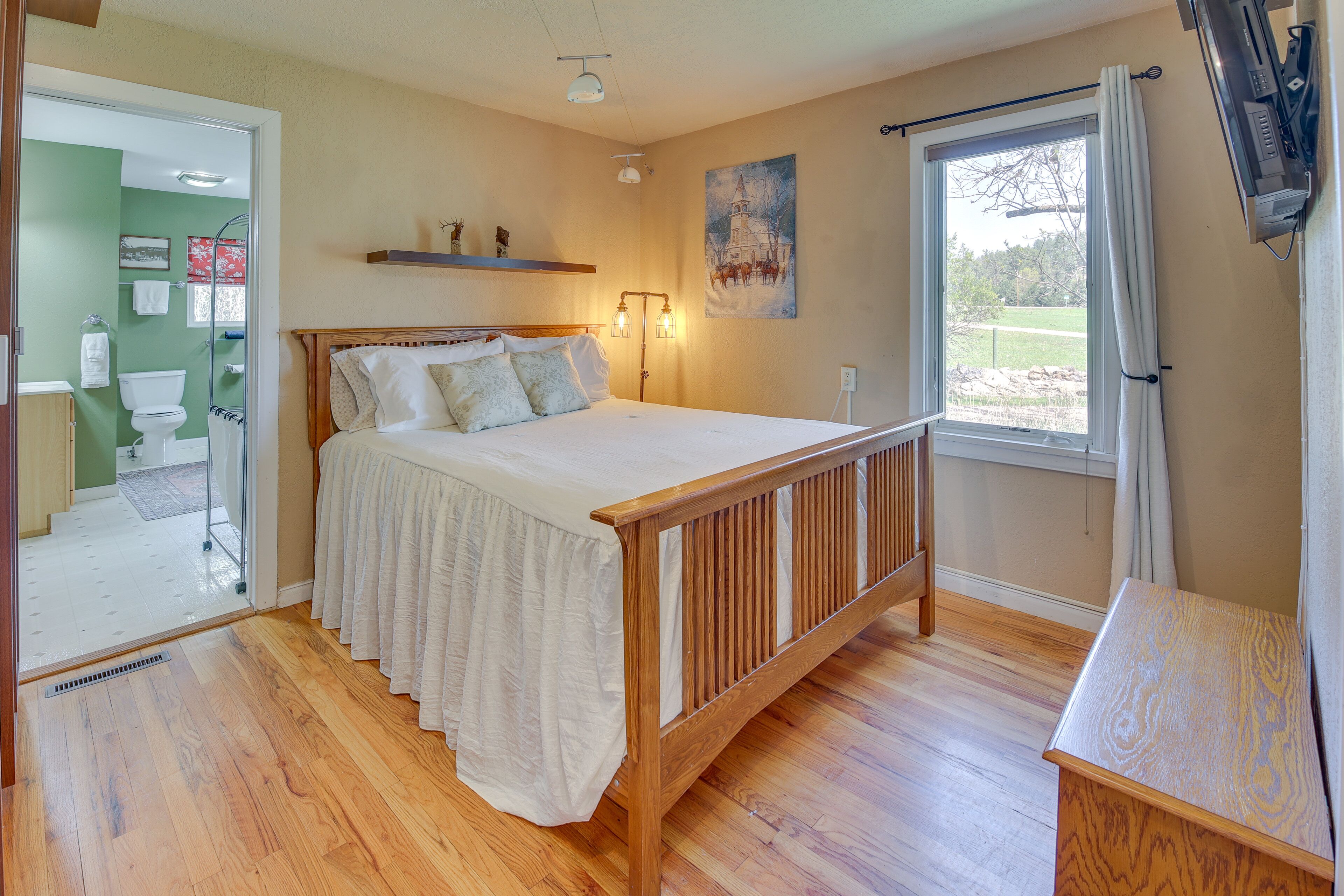 A bedroom with a wooden bed, a nightstand, a window with curtains, a painting on the wall, and a bathroom visible through an open door.