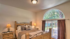 Un dormitorio con cama de madera, dos mesitas de noche con lámparas, un ventanal arqueado y vistas al verdor exterior.