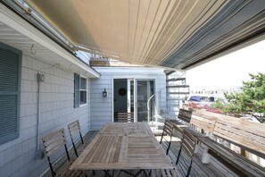 Een overdekt terras met een houten tafel en bankjes, een schuifpui naar een binnenruimte en uitzicht op de omgeving.