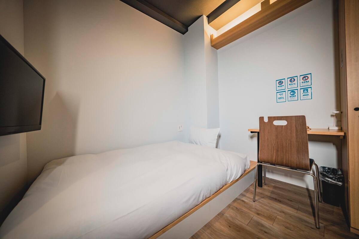 A small, neatly arranged bedroom with a single bed, a wooden chair, and a desk with a chair. There are wall-mounted light fixtures and a small trash bin.
