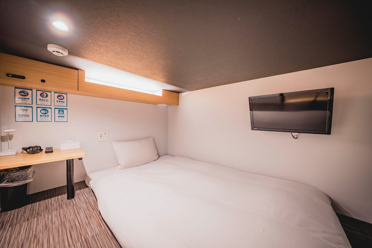 A hotel room with a bed, a desk with a chair, a television mounted on the wall, and a ceiling light.
