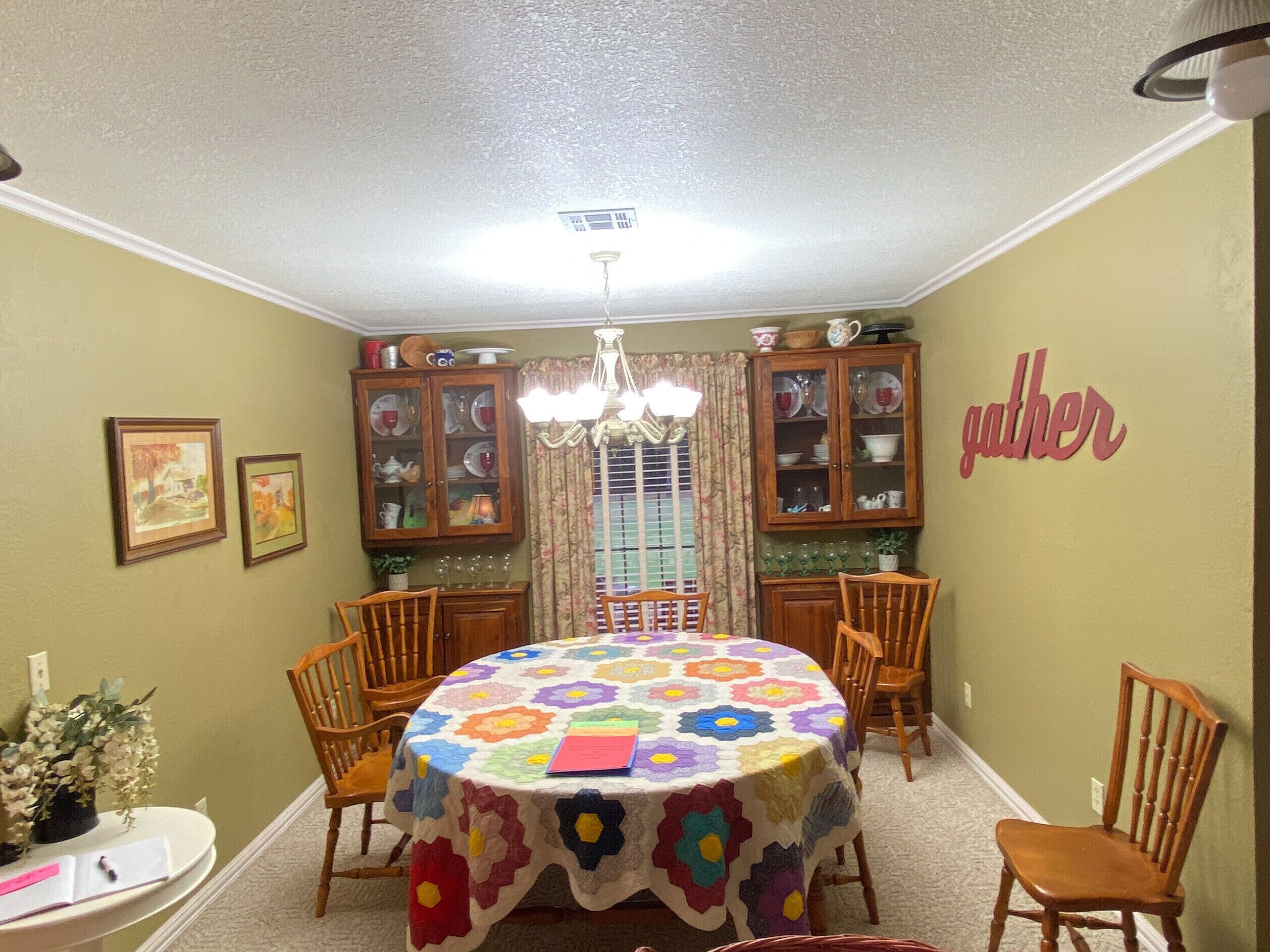 Comedor con una mesa redonda cubierta por un colcha de colores, sillas de madera a cada lado y un aparador con puertas de vidrio arriba.