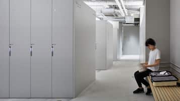 A modern hotel corridor with lockers numbered 551 to 559, a person sitting on a bench with a suitcase, and a concrete ceiling with exposed pipes.