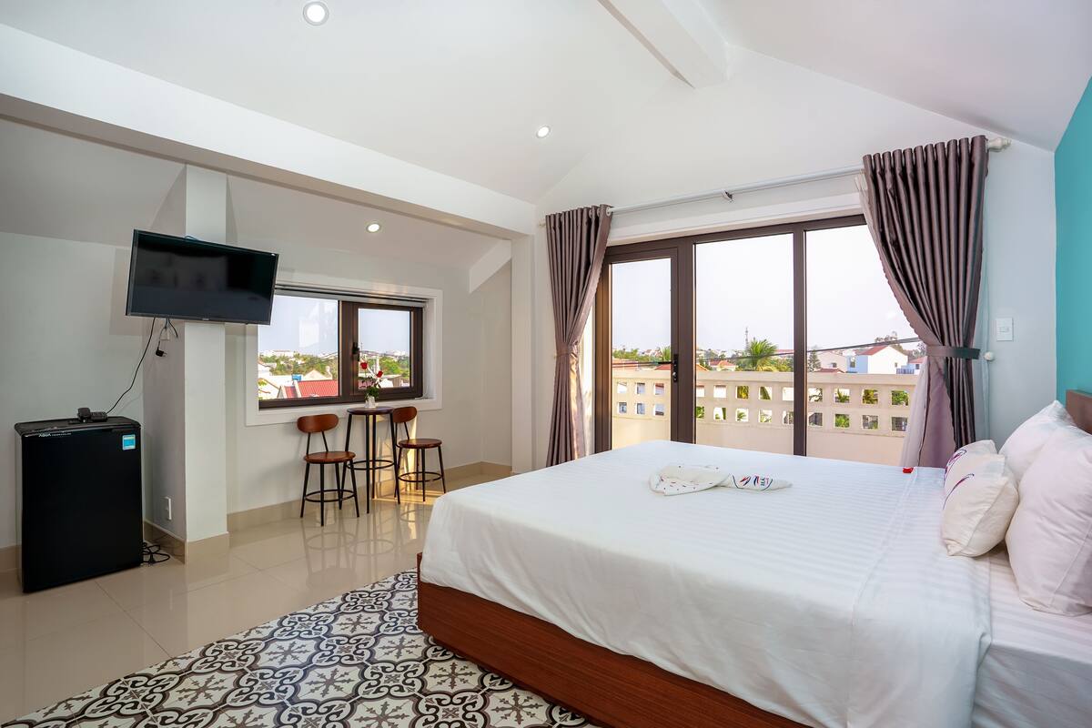 A hotel room with a large bed, a TV mounted on the wall, a small desk with chairs, and a window with curtains.