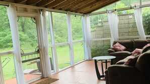 Eine überdachte Terrasse mit Holzboden, großen Fenstern, einem braunen Sofa und einem kleinen runden Tisch.