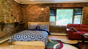 Ein Schlafzimmer mit einer Ziegelwand, einem Bett mit gemusterten Betttextilien, einem roten Ledersofa und einem Fenster mit Jalousien.