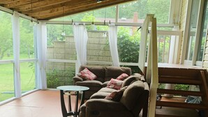 Eine überdachte Veranda mit Holzboden, eine gepolsterte Sitzecke mit einem Tisch und Blick auf einen grünen Garten durch große Fenster.