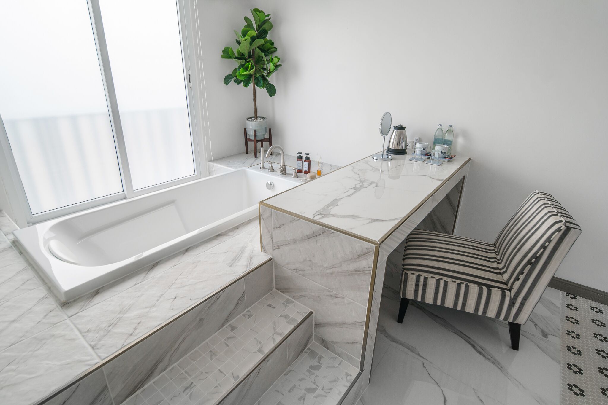 Un baño moderno con una bañera grande, un tocador de mármol, una silla a rayas y una planta en maceta.