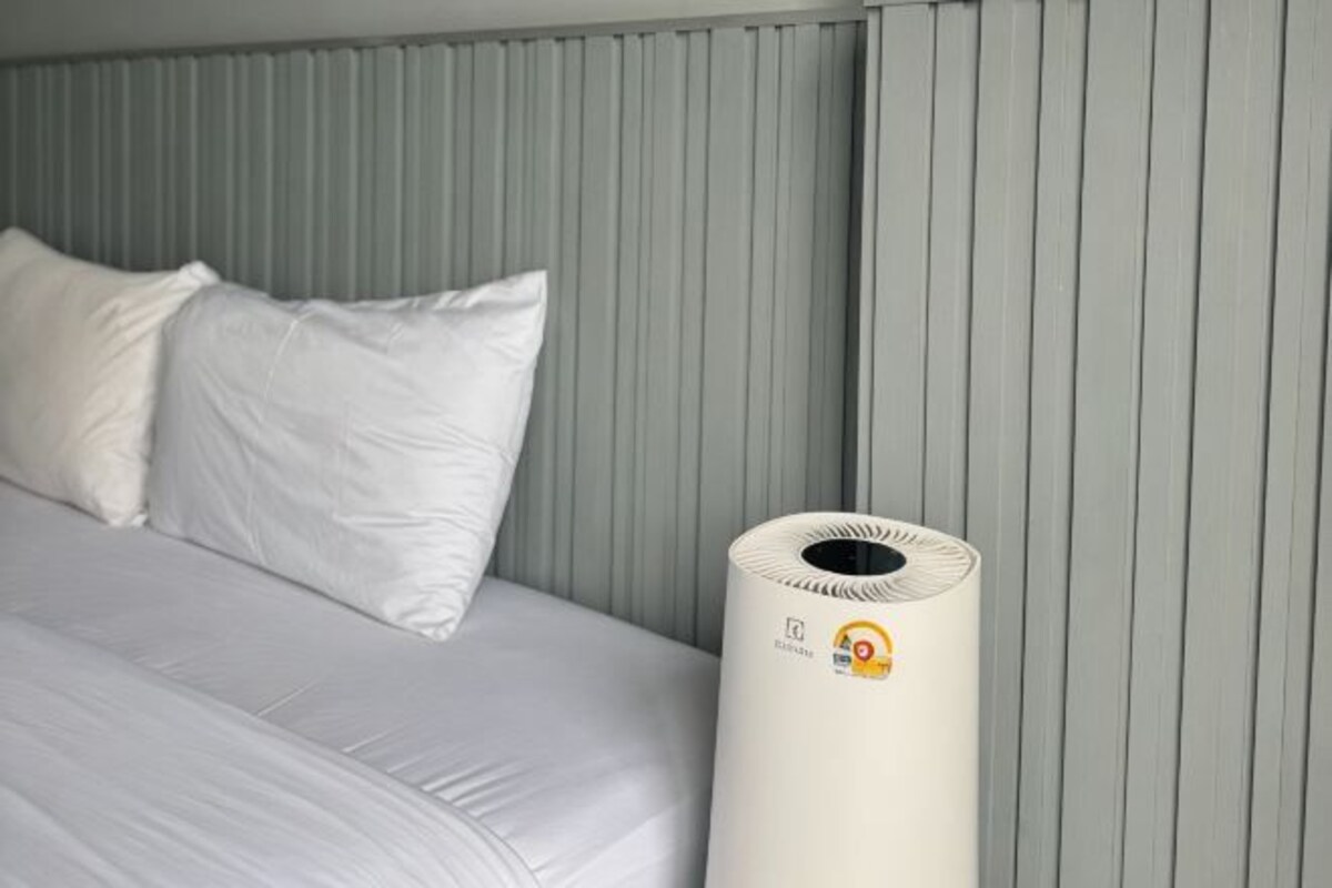 A bed with white bedding and pillows, a white cylindrical object, and a grey wall with vertical lines.