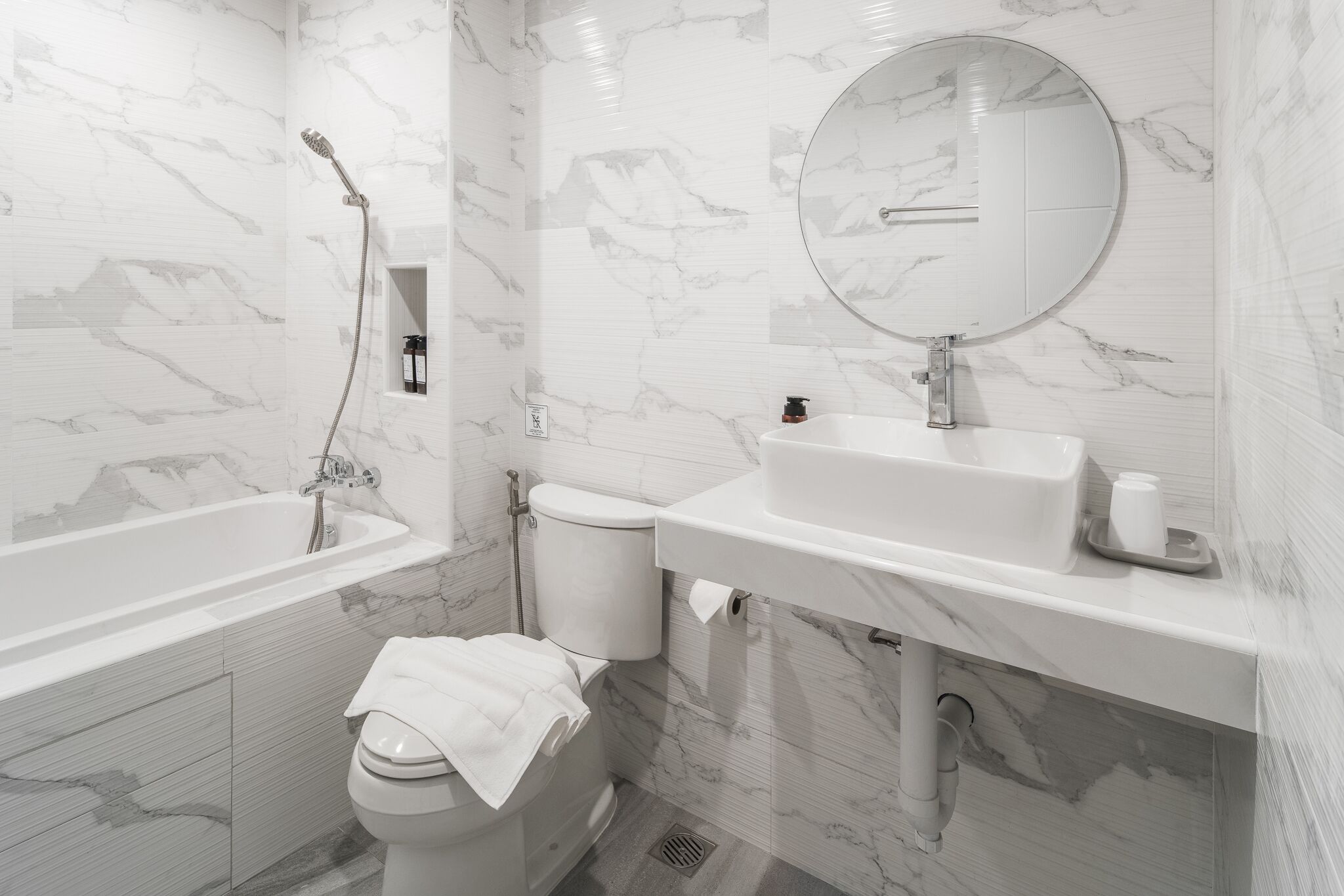 A modern bathroom with a marble backsplash, a round mirror, a bathtub, and a toilet.