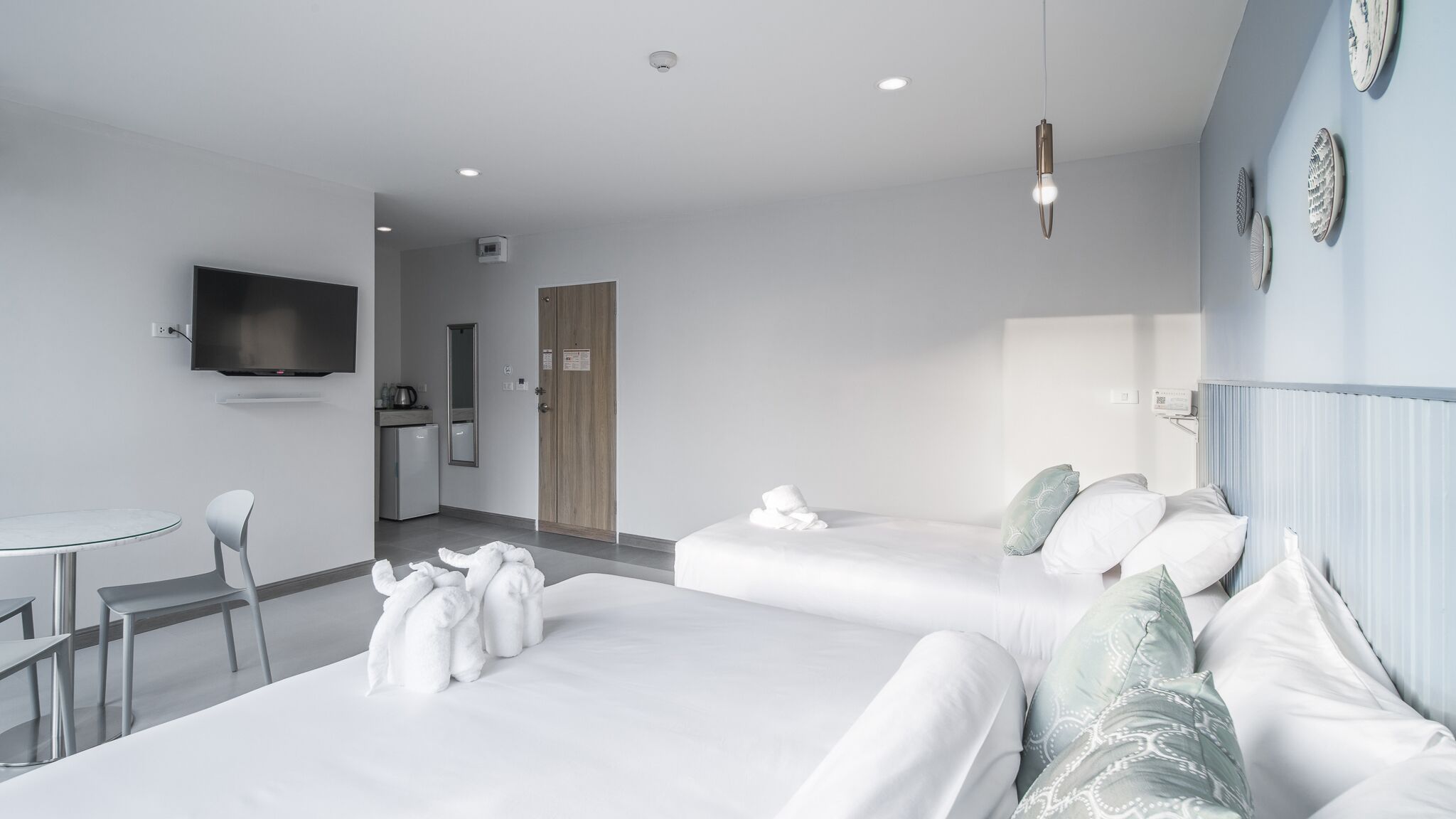 A modern hotel room with two beds, a small dining area, a TV, and a kitchenette.
