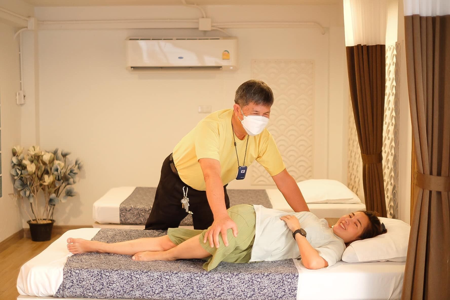 A medical examination room with a patient lying on a bed, a healthcare professional attending to them, and an air conditioning unit on the wall.