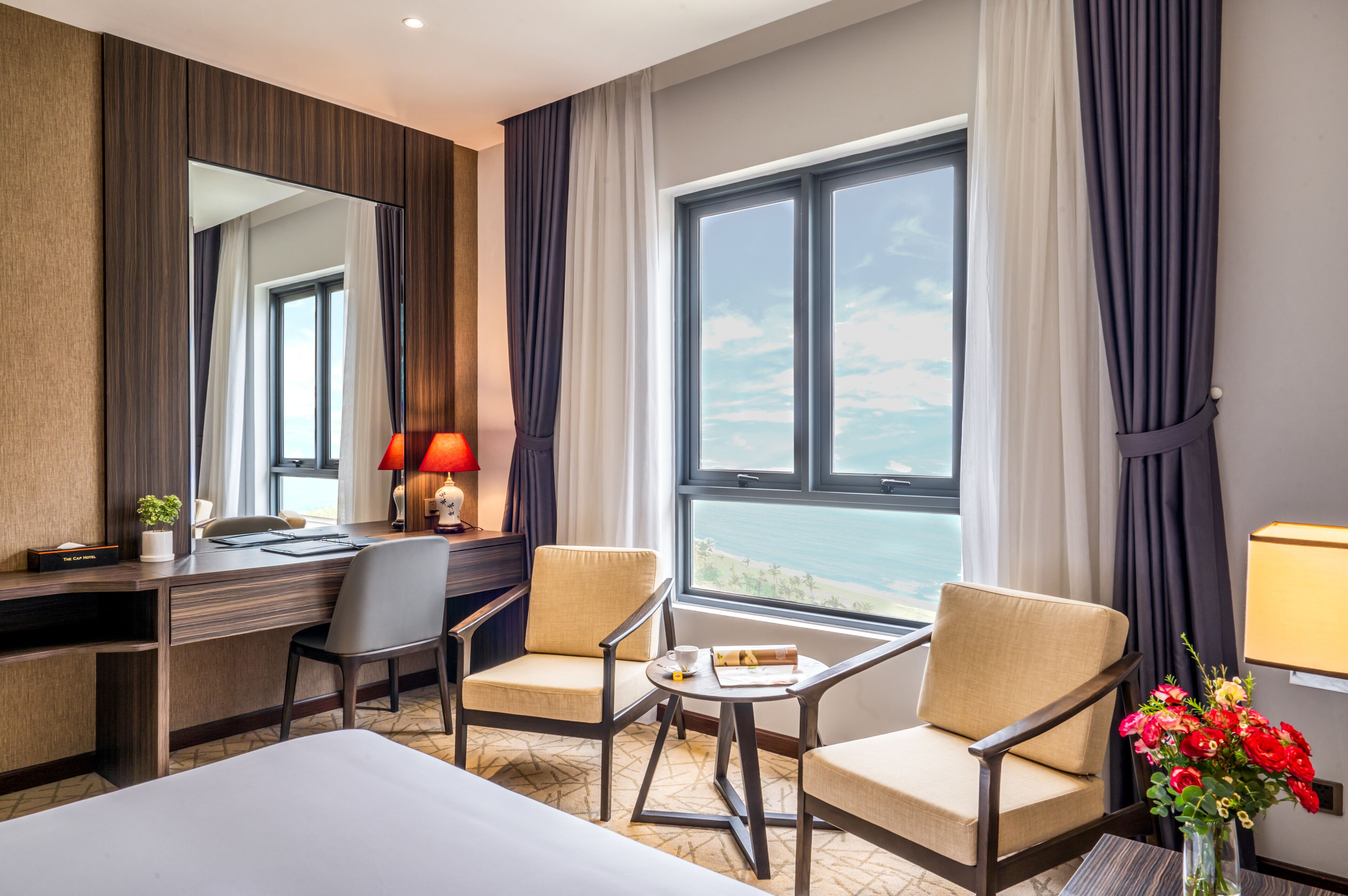 A hotel room with a large window, a desk, two chairs, a small table, and a bed.