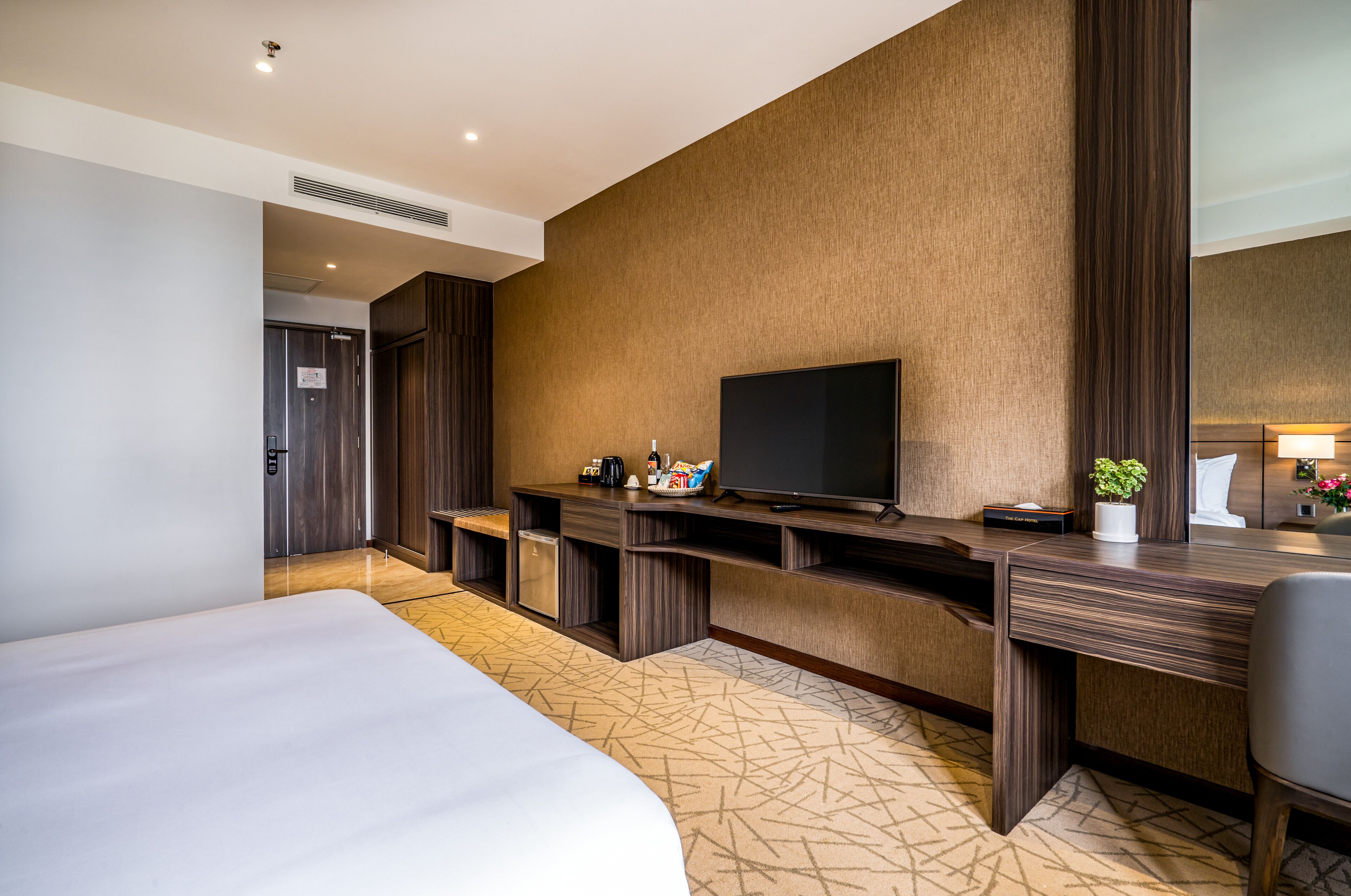 A hotel room with a large bed, a desk with a flat-screen TV, a small coffee maker, and a potted plant.