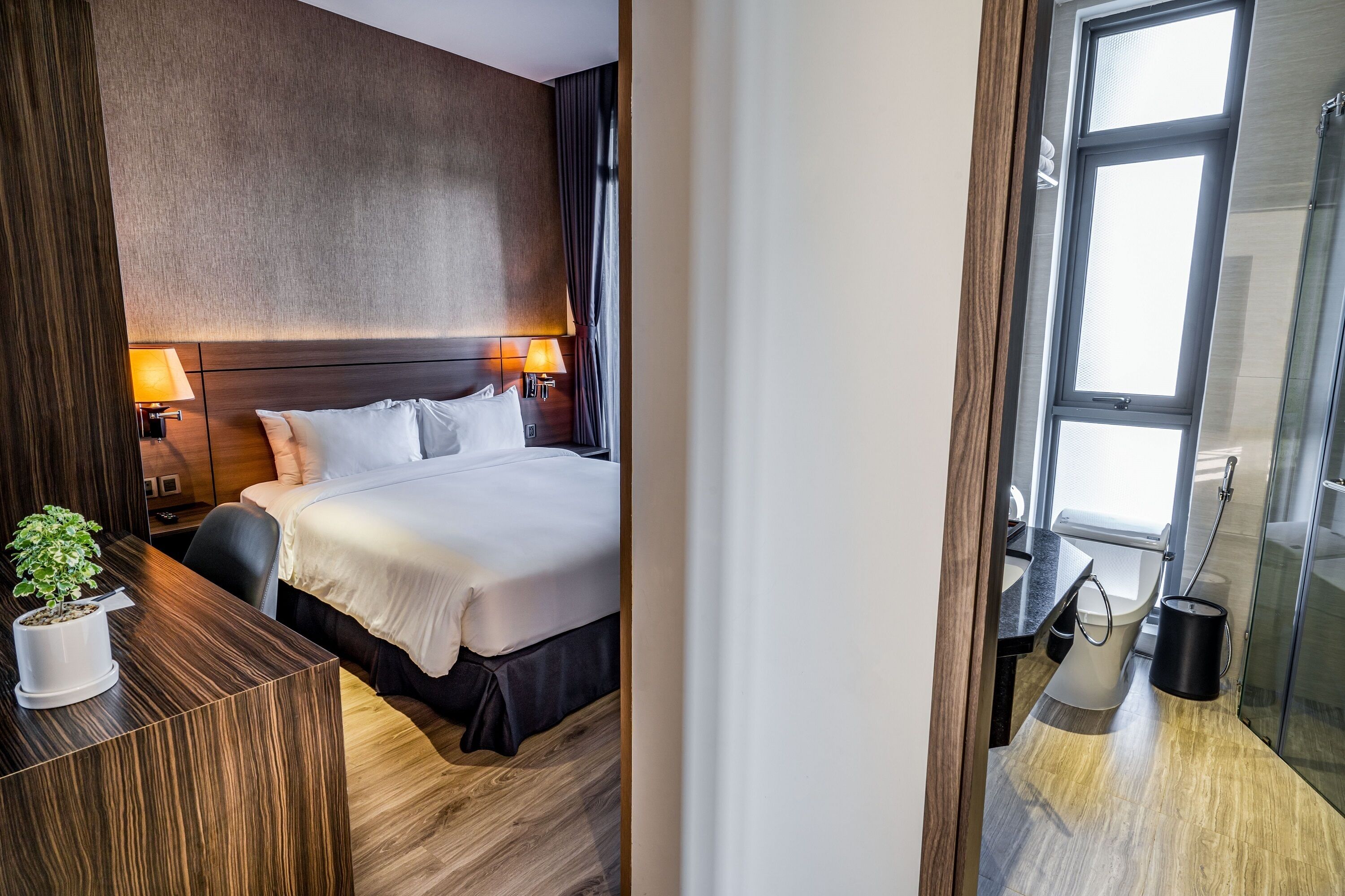 A hotel room with a large bed, two bedside lamps, a wooden headboard, a small potted plant, and a view of the bathroom through a glass door.