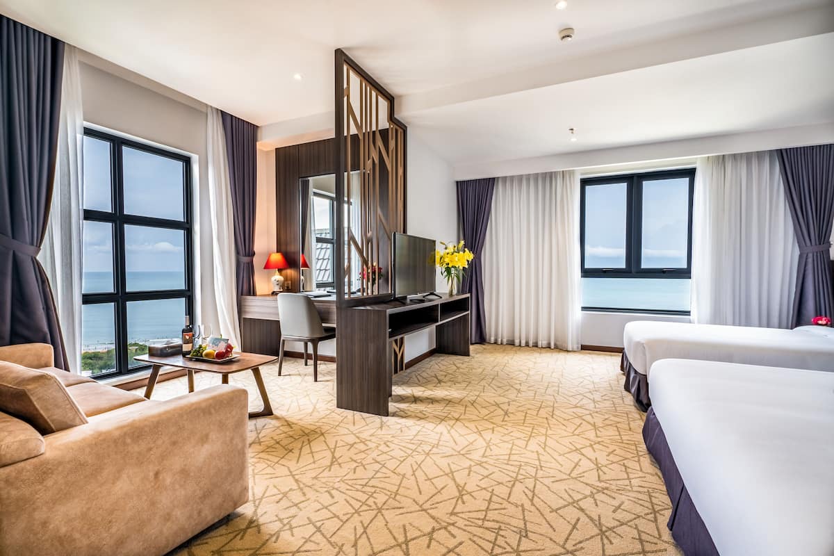 Quarto de hotel com uma cama grande, uma escrivaninha com televisão, um sofá e vista para o mar.