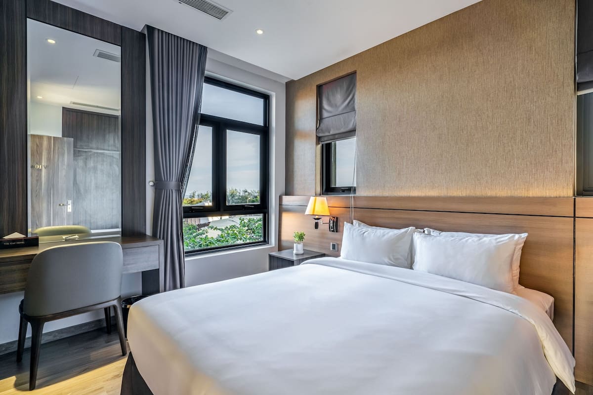 Quarto de hotel com uma cama grande, uma mesa com cadeira, uma janela com cortinas e uma mesa de cabeceira com luminária.