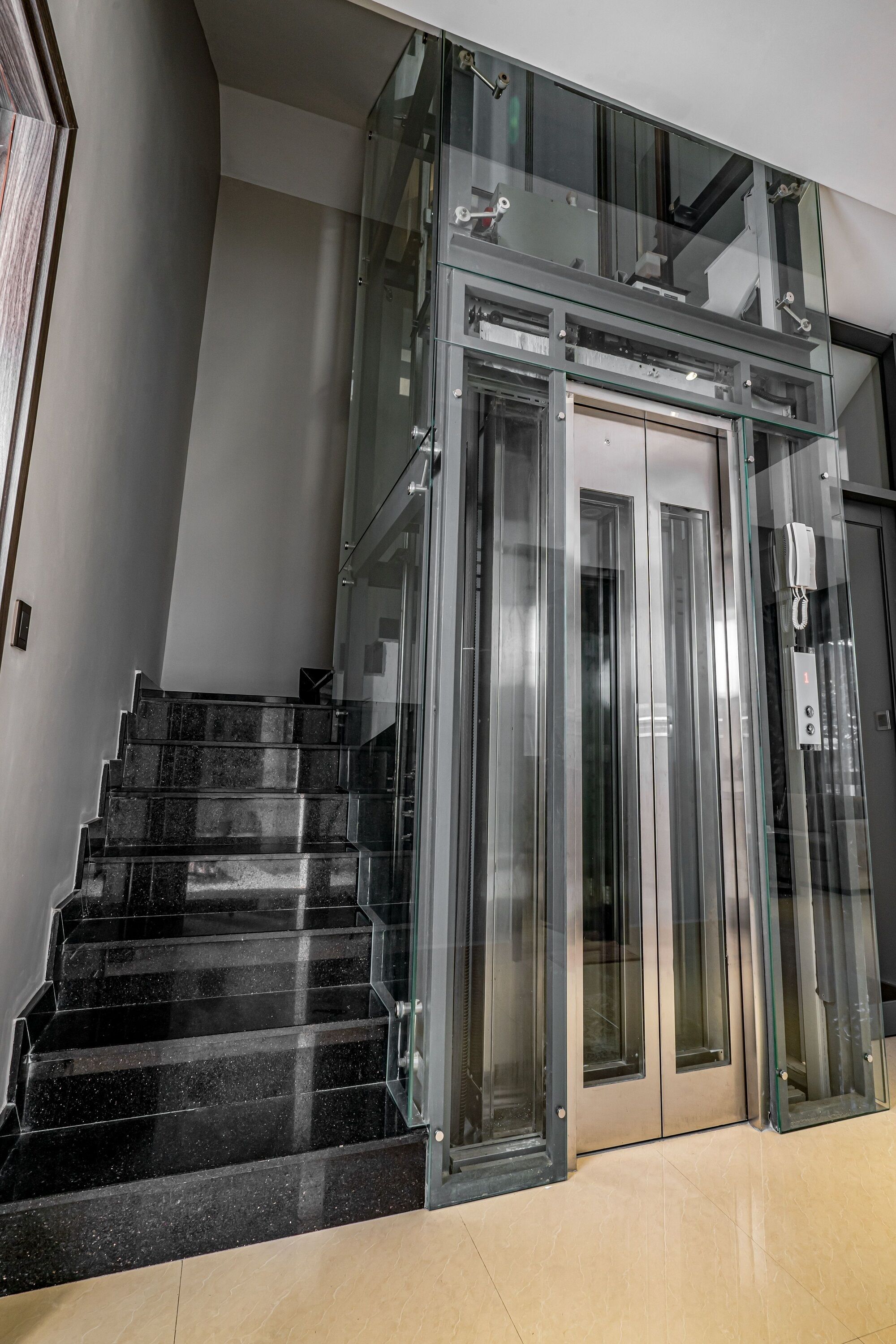 A modern elevator with glass doors and a metal frame, next to a staircase with black marble steps.