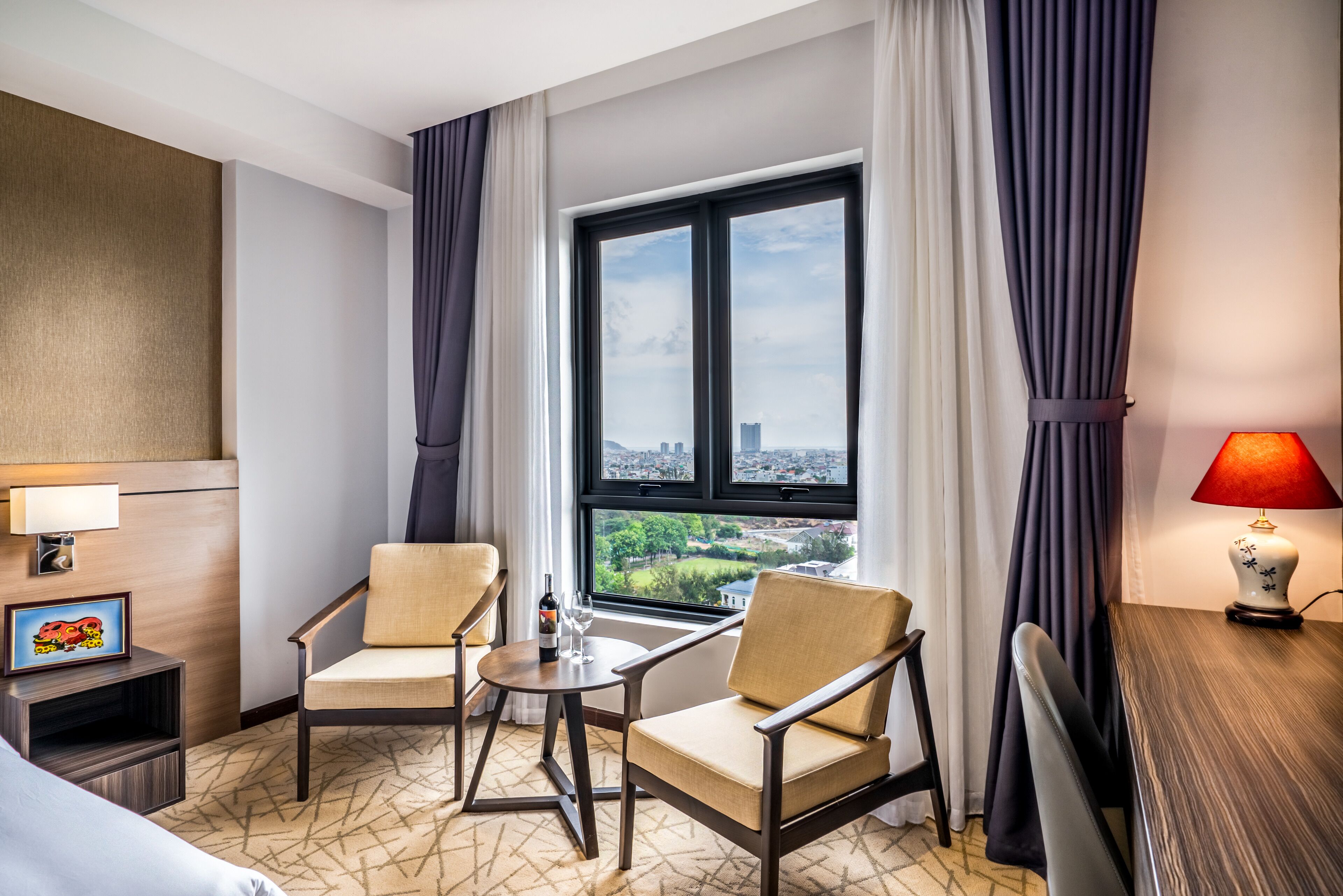 A hotel room with two armchairs, a small table with a bottle and glass, a lamp, and a view of the city through a large window.