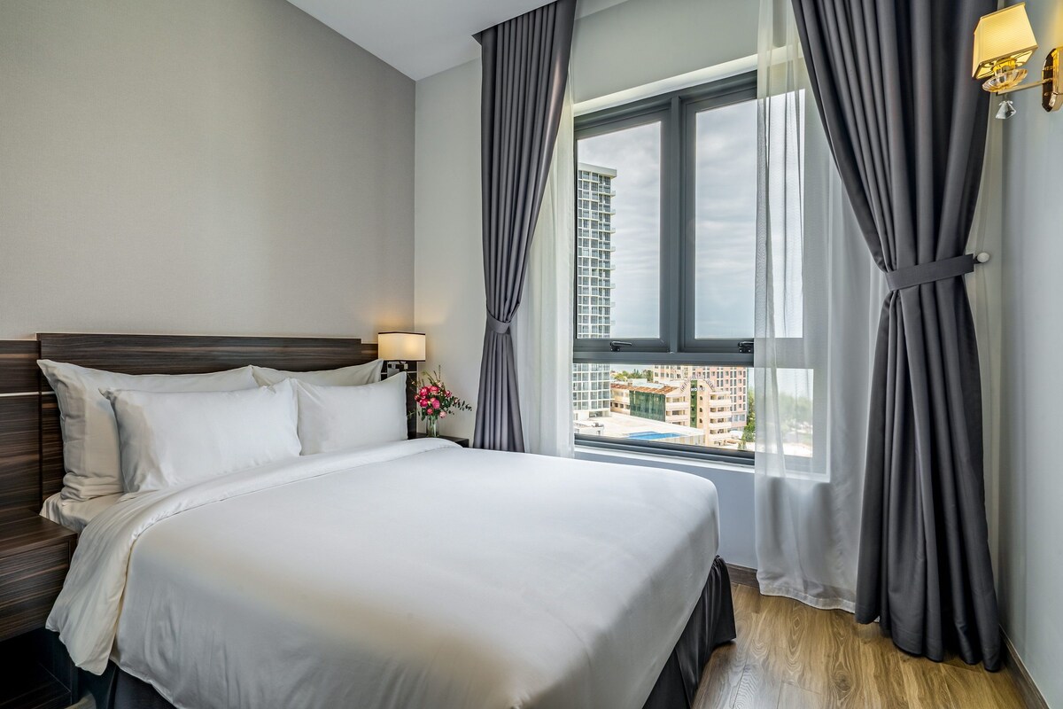 A hotel room with a large bed, a nightstand with a lamp, and a view of a cityscape through a window with curtains.