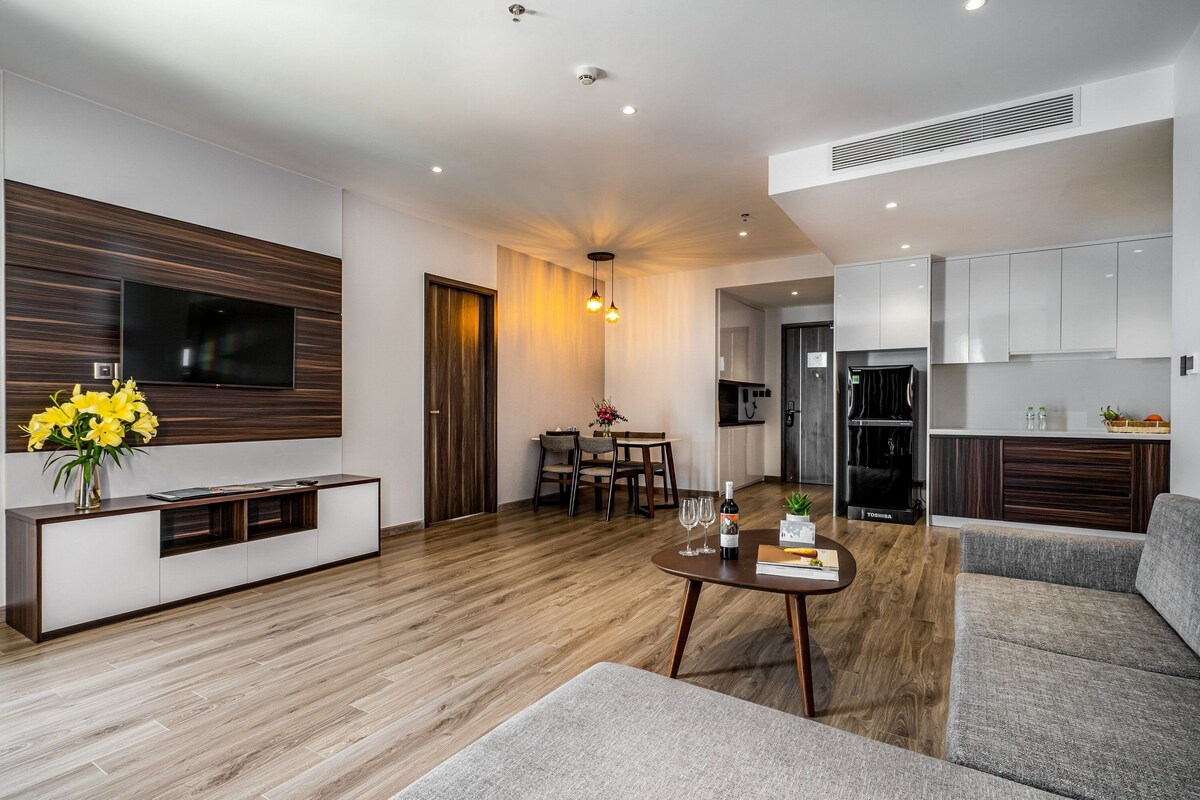 Una sala moderna con televisor de pantalla plana, piso de madera, comedor con mesa y sillas, y cocina con armarios blancos y refrigerador negro.