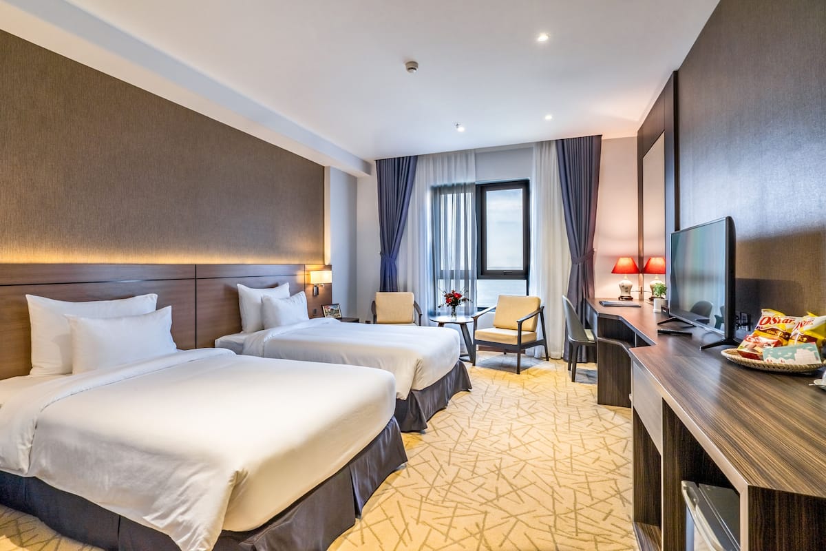 Quarto de hotel com duas camas, uma escrivaninha com televisão, uma cadeira e uma janela com cortinas.