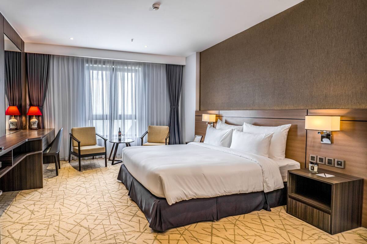 Quarto de hotel com uma cama grande, duas mesas de cabeceira com abajures, uma escrivaninha com duas cadeiras e uma janela ampla com cortinas.