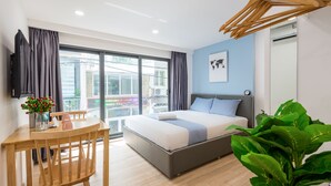 A modern bedroom with a bed, a desk, and a chair. There is a large window, a plant, and a wall-mounted TV. - Cozrum Homes Saphera Residence (Ho Chi Minh City)
