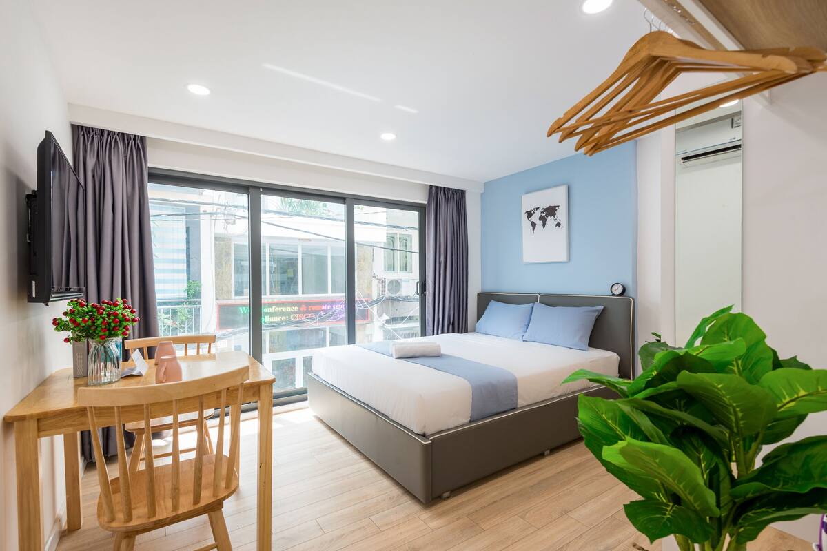 Un dormitorio moderno con cama, escritorio y silla. Hay un ventanal grande, una planta y un televisor montado en la pared.