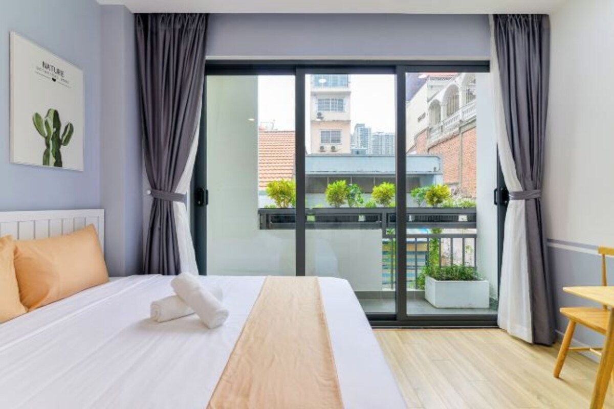 A hotel room with a bed, a small table, a window with curtains, and a balcony with plants.