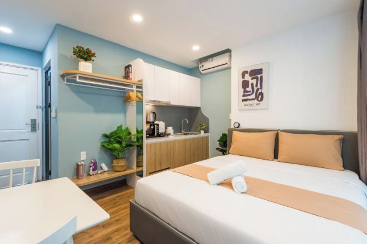 A hotel room with a bed, a small kitchenette, a white table, and a shelf with plants and toiletries.