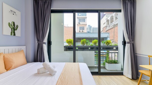 A modern hotel room with a large bed, a view of the city through a sliding glass door, and a small balcony with plants. - Cozrum Homes Saphera Residence (Ho Chi Minh City)