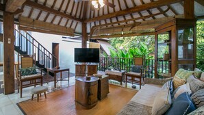 A spacious living area with a wooden ceiling and a television. There are wooden furniture pieces, including a sofa and chairs, and a large window offering a view of greenery. - Villa D'Kali 999 (Seminyak)