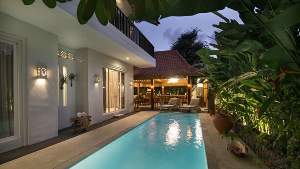 A swimming pool with a covered patio area, surrounded by lush greenery and a modern building with a red-tiled roof. - Villa D'Kali 999 (Seminyak)