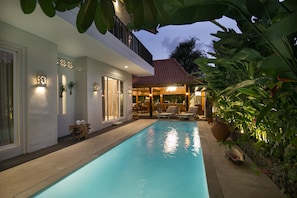 A swimming pool with a covered patio area, surrounded by lush greenery and a modern building with a red-tiled roof. - Villa D'Kali 999 (Seminyak)