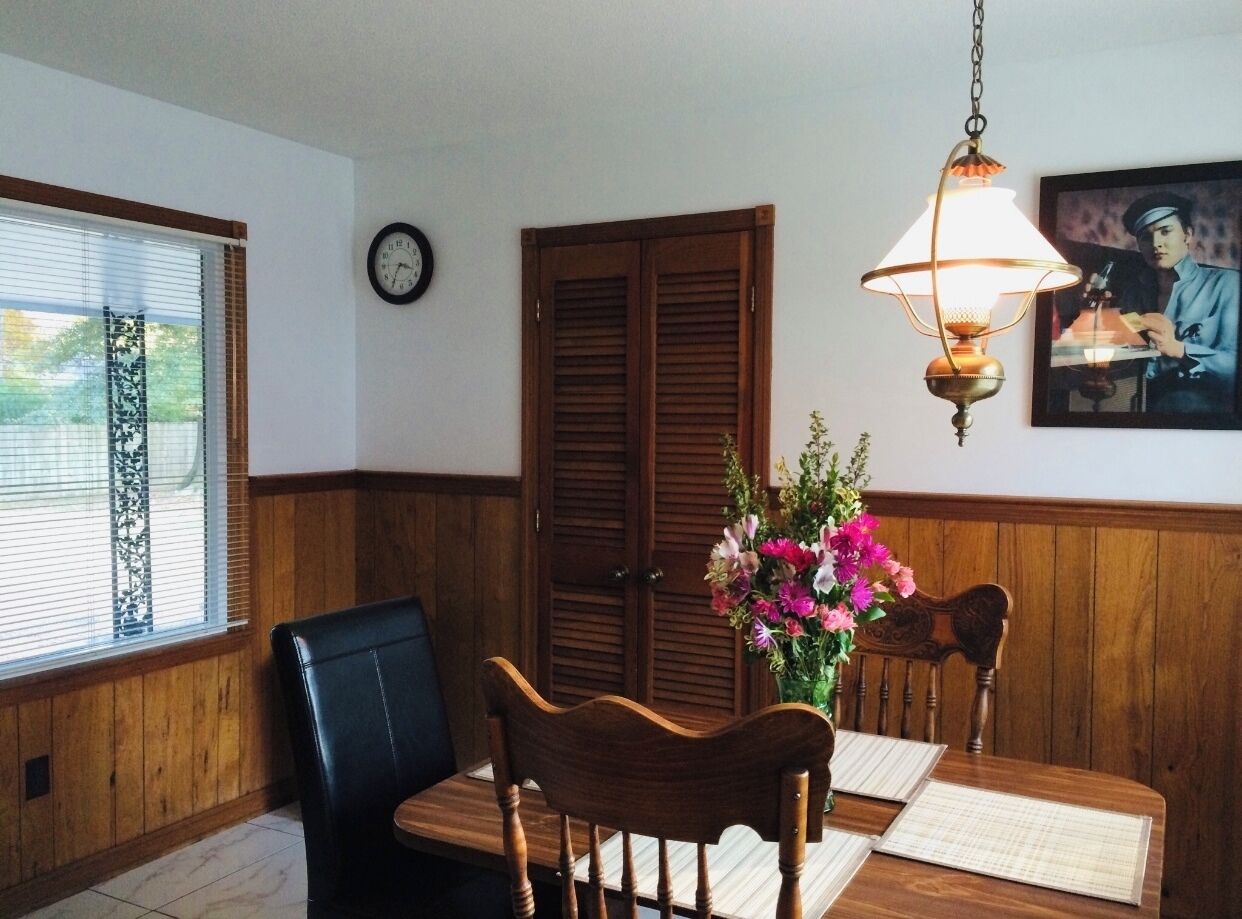 Un coin repas avec une table en bois, deux chaises, un vase de fleurs, une horloge au mur et un tableau encadré.