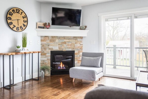 A living room with a fireplace, a grey armchair, a wooden coffee table, and a TV mounted above the fireplace. - Hygge | 3BD Townhome | Downtown | Wineries | Beaches (Traverse City)
