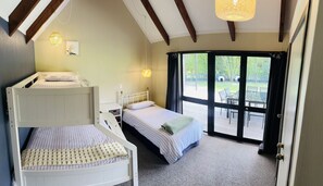 A bedroom with bunk beds, a single bed, a small bedside table, a lamp, and a sliding glass door to an outdoor area. - Private family friendly slice of paradise- ready for you to enjoy. (Mahia)