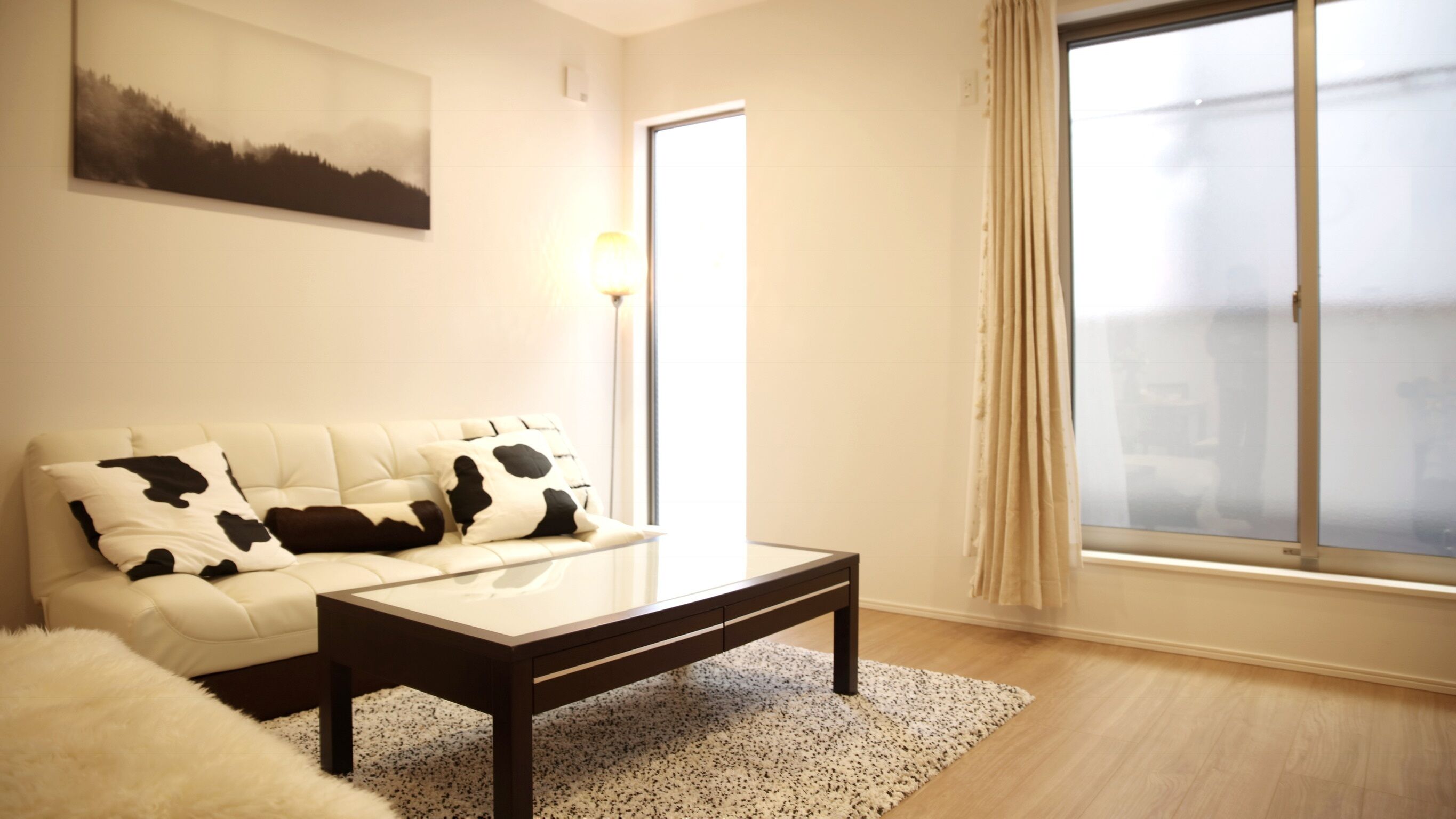 A living room with a white sofa, a coffee table, a rug, and a large window with curtains.