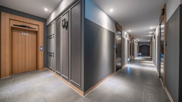 Un pasillo moderno de hotel con una puerta de madera a la izquierda, un ascensor grande y un largo pasillo con puertas a ambos lados.