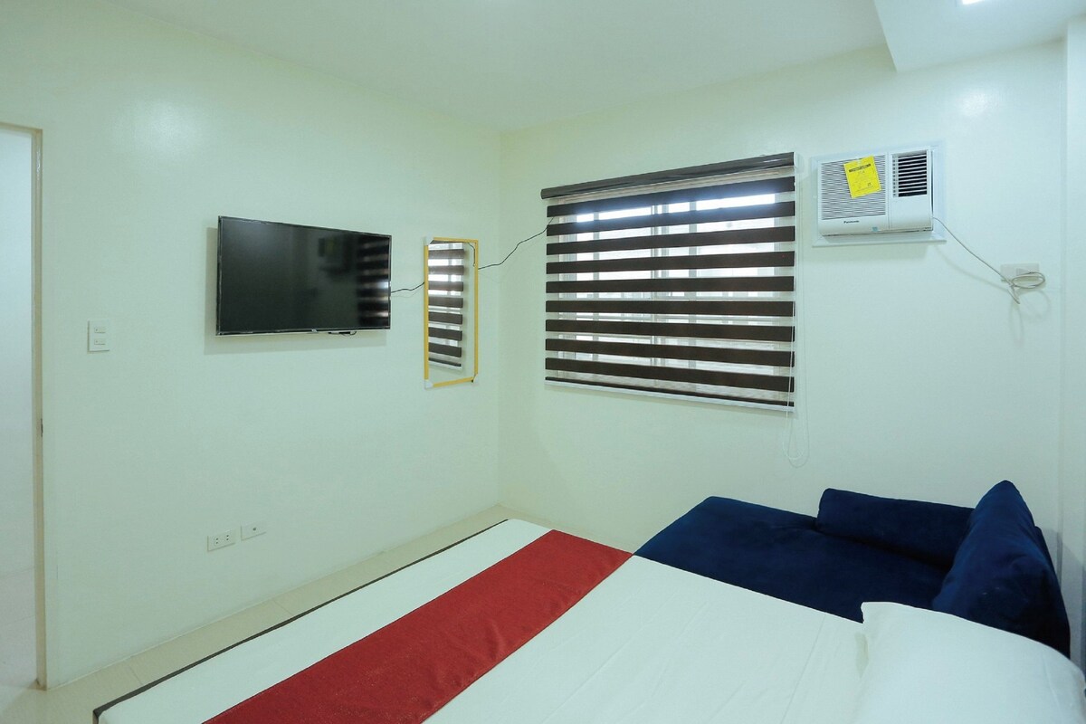 A room with a bed, a TV, a window with blinds, and an air conditioning unit.