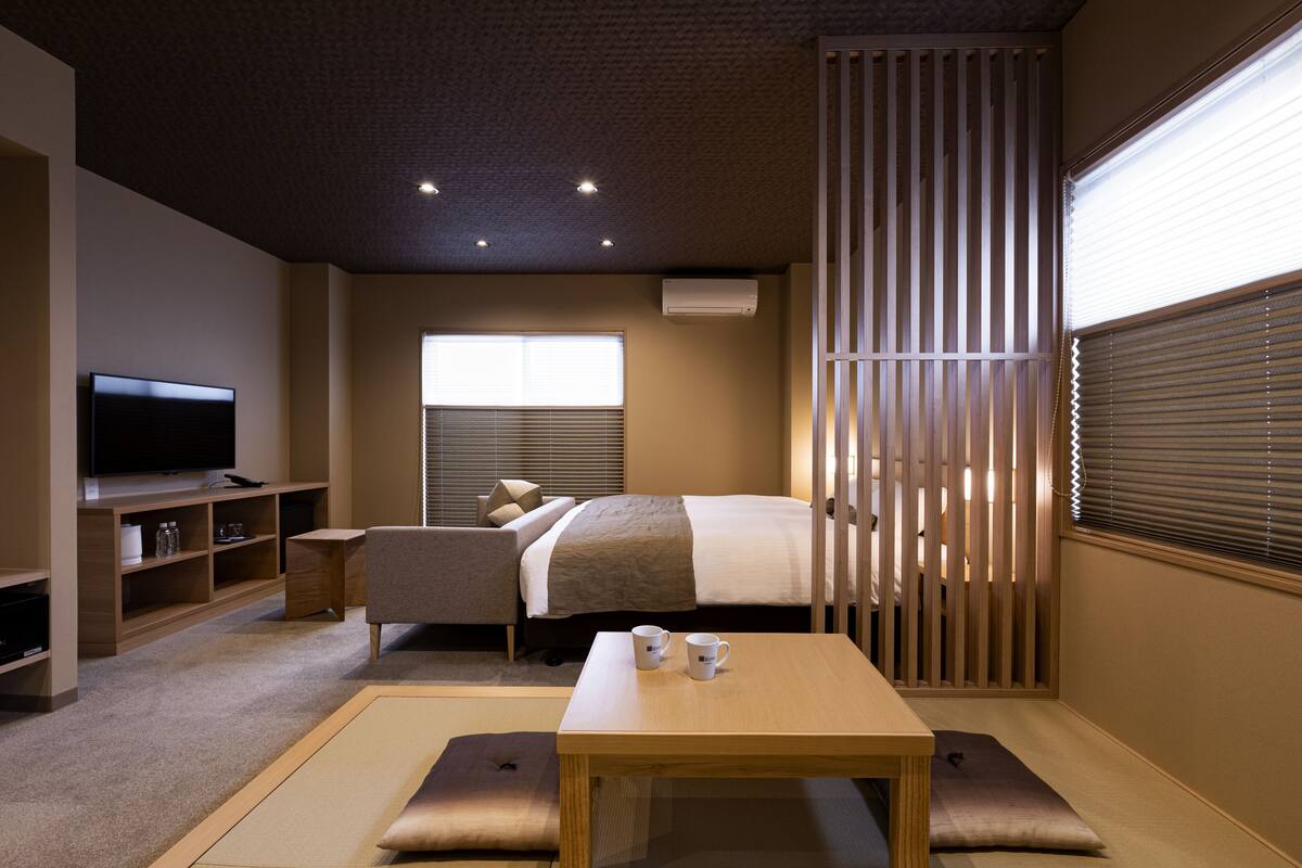 Habitació d'hotel moderna amb televisió de pantalla plana, un llit amb una finestra gran, una tauleta de nit amb dues tasses i una porta corredissa japonesa tradicional.