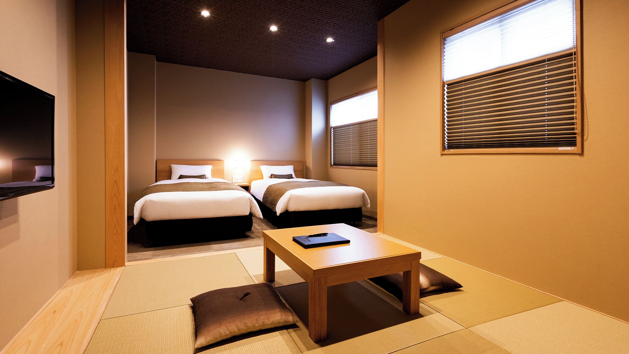 A hotel room with two beds, a flat-screen TV, a wooden table, and tatami mats.