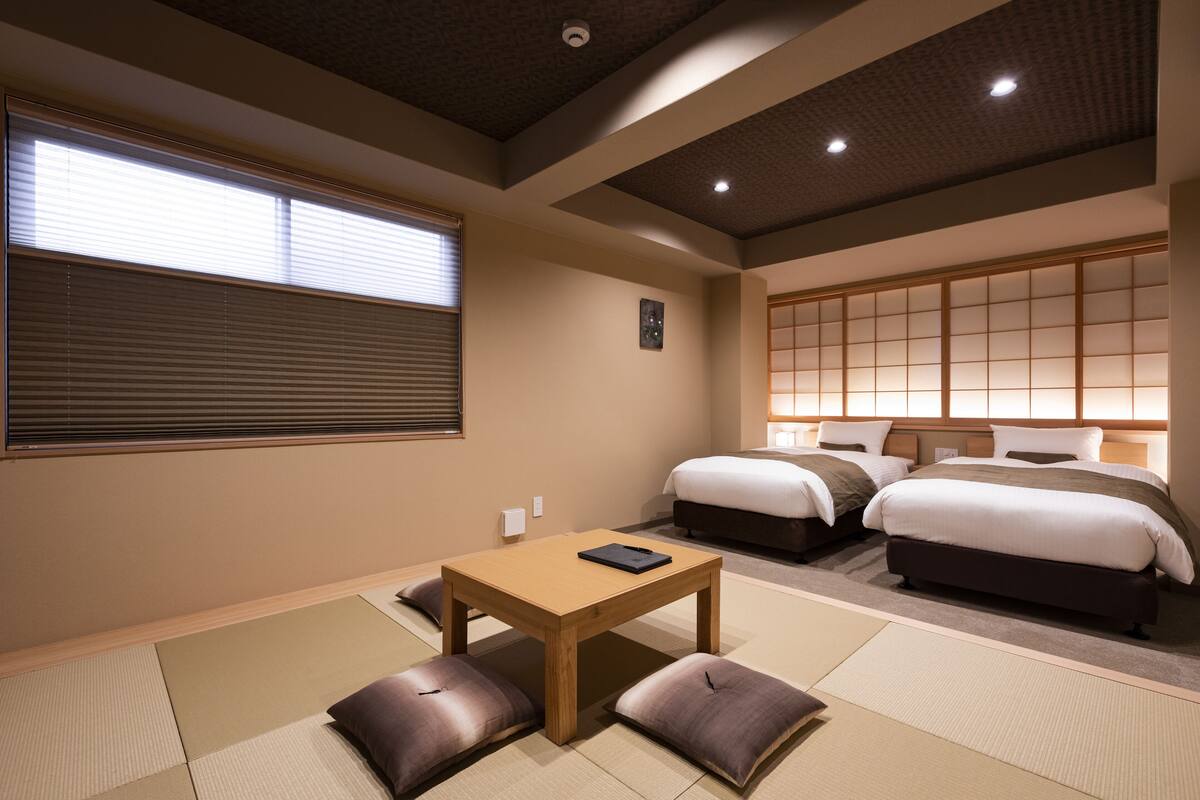 Habitació tradicional japonesa amb fons de tatami, taula de fusta baixa, dos llits amb roba de llit fosca i un panell corredís shoji.