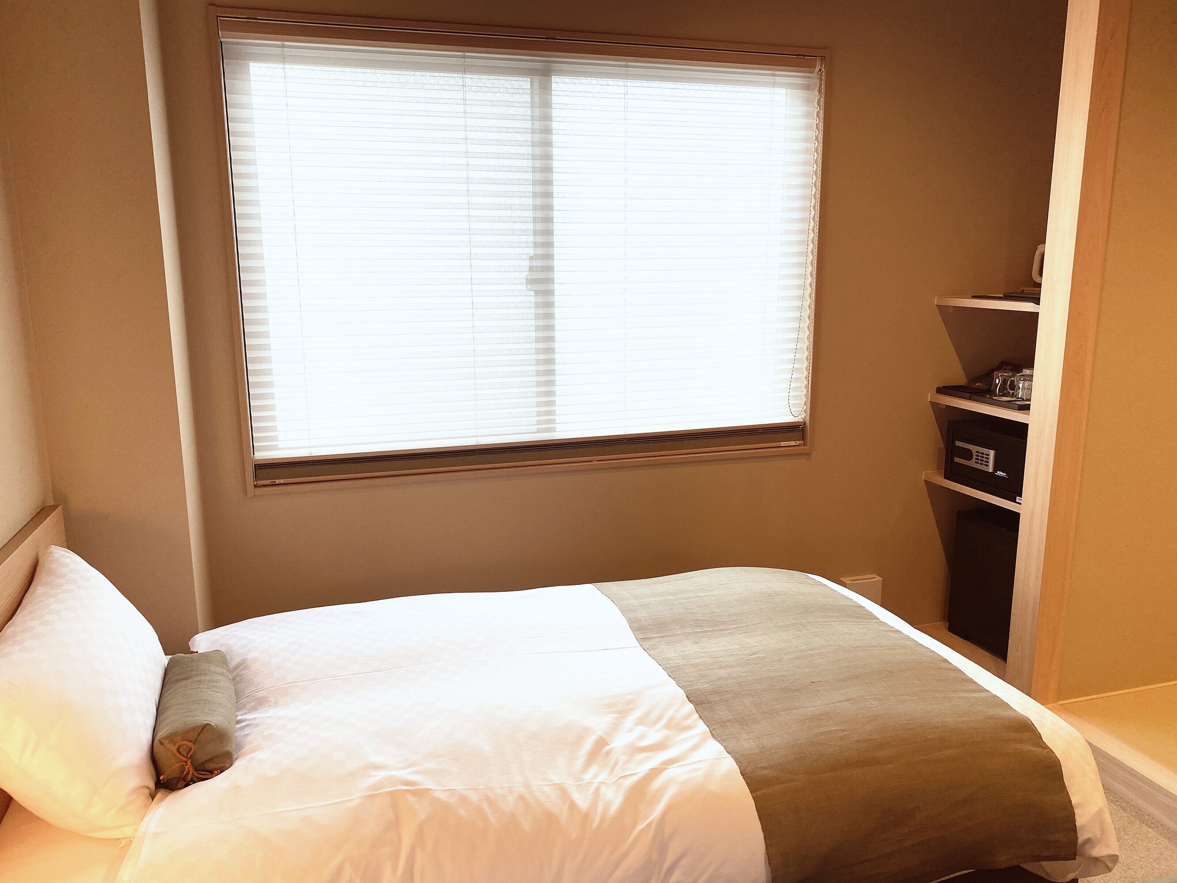 A bedroom with a bed, a window with blinds, and a shelf with items.