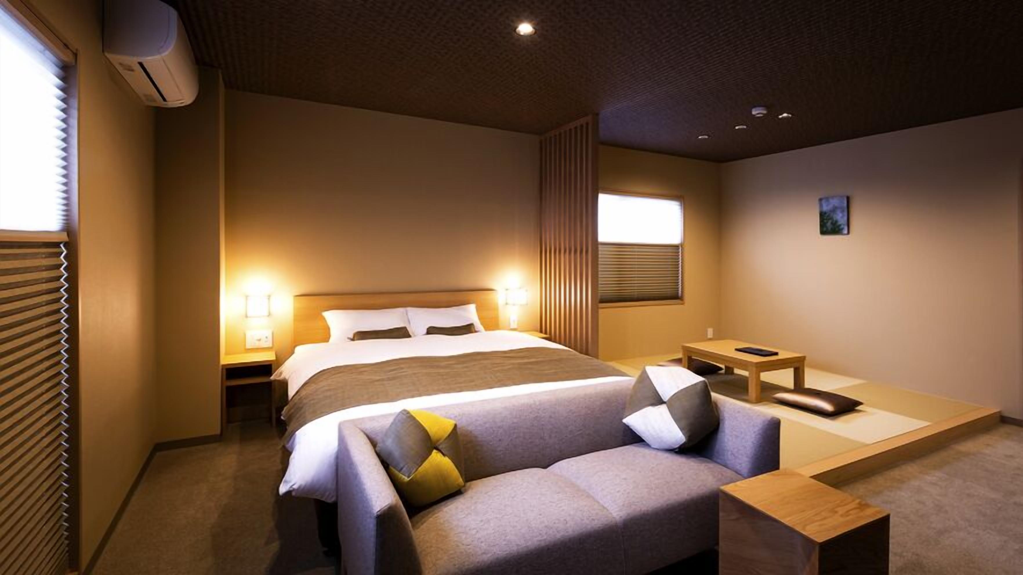 A modern hotel room with a large bed, bedside lamps, a sofa, a small table, and a window with blinds.